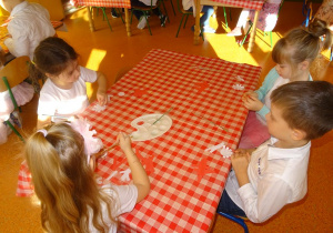 Czwórka dzieci przygotowuje kotylion z pasków papieru, sklejają elemnty.